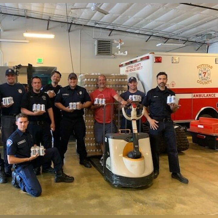A group of people in a garage-like setting holding cans, posing in front of a pallet and an emergency vehicle.