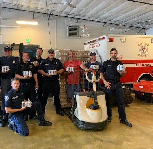 A group of people in a garage-like setting holding cans, posing in front of a pallet and an emergency vehicle.