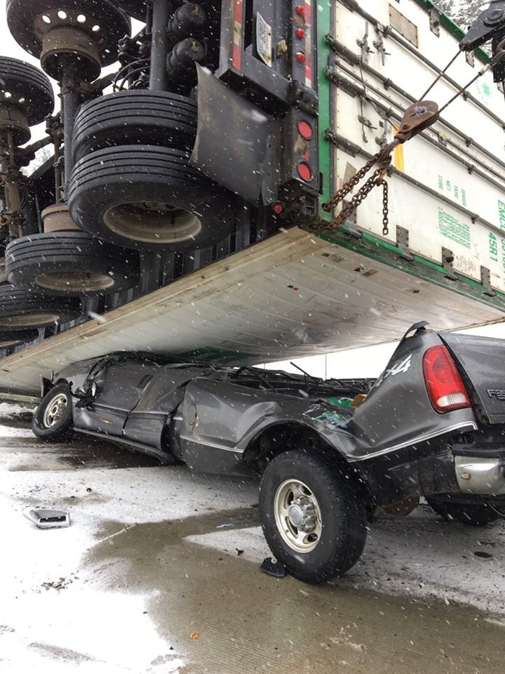 A truck is crushed under an overturned trailer in snowy conditions.