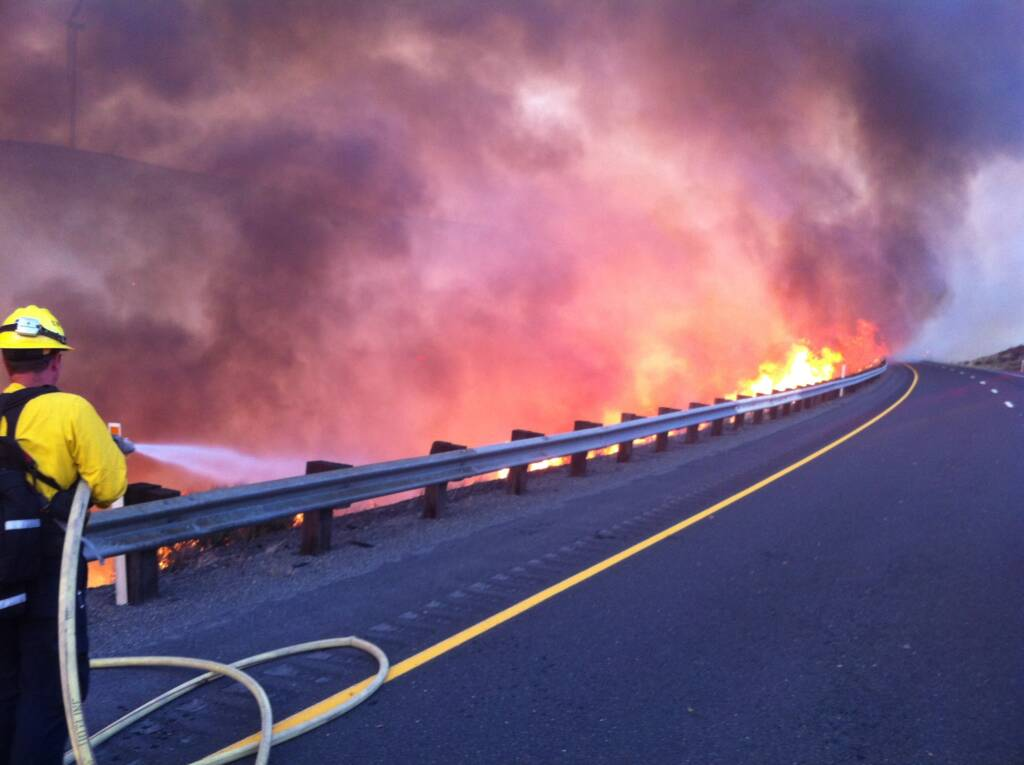 A firefighter battles a roadside blaze with heavy smoke on a highway.