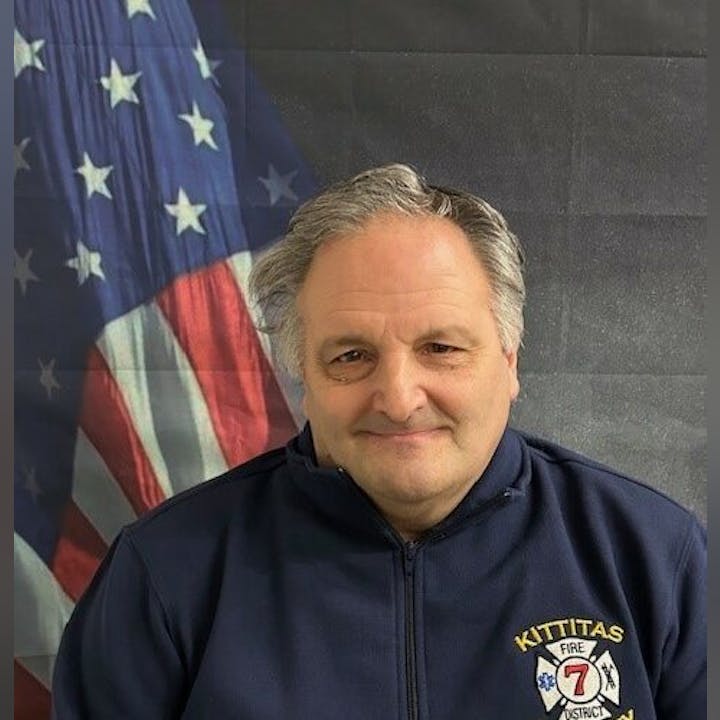 A person in a "Kittitas Fire District 7" jacket stands before a U.S. flag background.