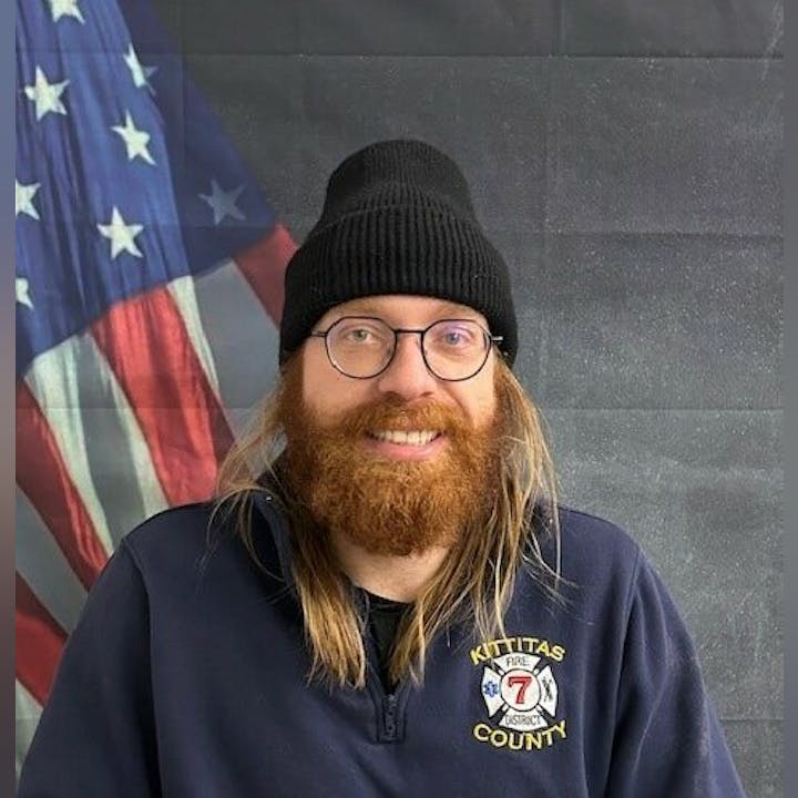 A person with glasses and a beanie, wearing a "Kittitas County Fire District 7" jacket, stands in front of a U.S. flag backdrop.