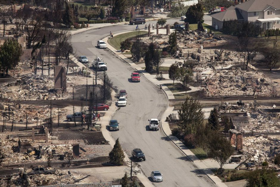 Aerial view of a neighborhood with severe fire damage, showing burned houses and charred trees along a street with some cars.