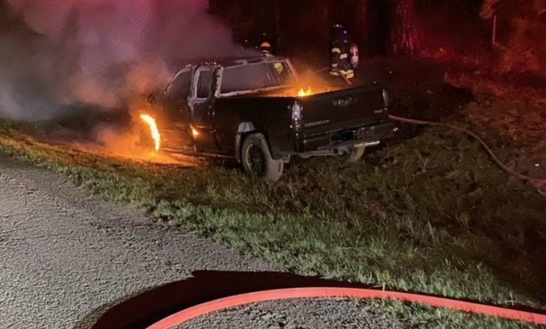 A black pickup truck is on fire by the roadside, with firefighters present and a hose nearby.