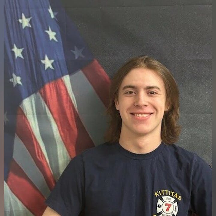 A person smiling, wearing a "Kittitas" t-shirt, standing in front of an American flag backdrop.