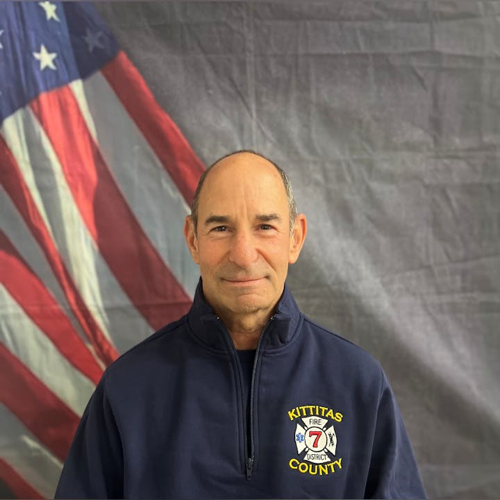 A person in a "Kittitas County Fire District 7" jacket stands before a U.S. flag backdrop.