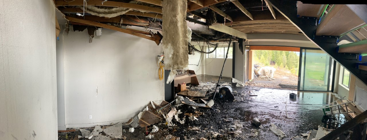 A damaged room with exposed beams, debris, and water on the floor, looking out to a grassy landscape.