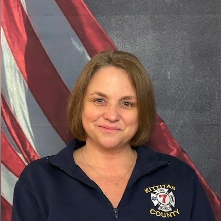 A person is smiling in front of an American flag, wearing a navy blue jacket with "Kittitas County" and a fire department emblem.