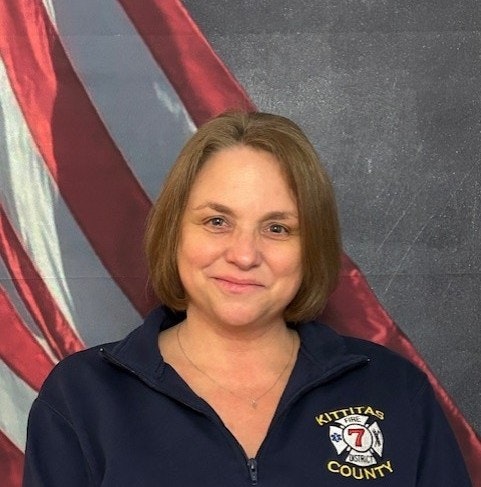 A person is smiling in front of an American flag, wearing a navy blue jacket with "Kittitas County" and a fire department emblem.