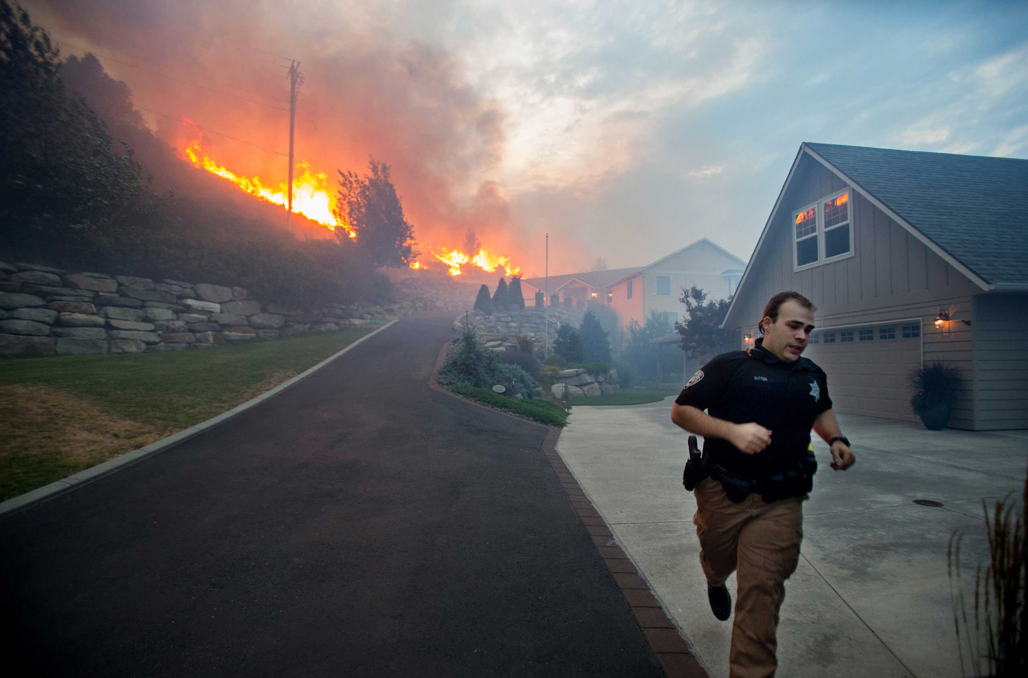 A person in uniform runs down a driveway as a hillside fire burns nearby, with smoke and flames visible.