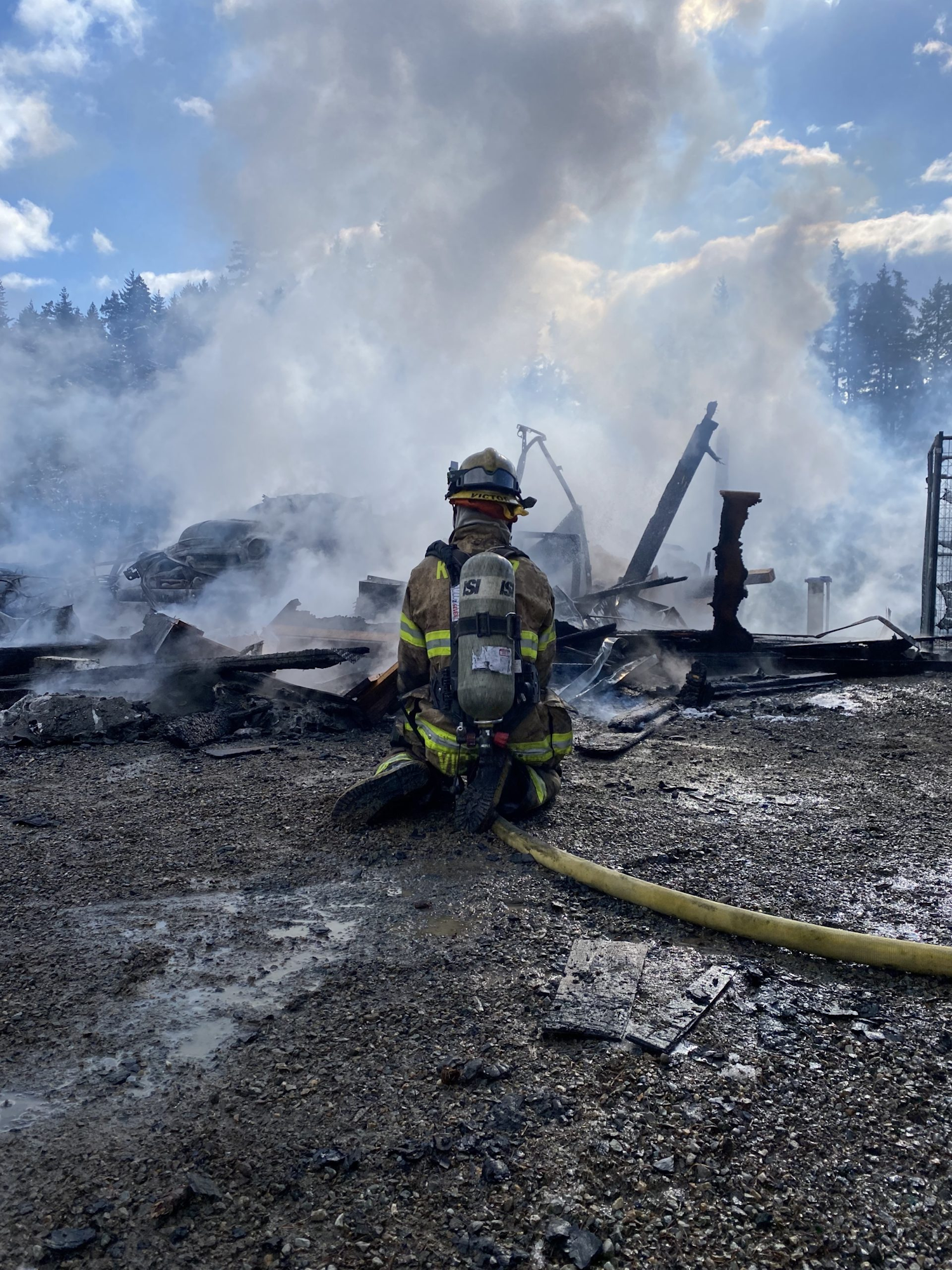 A firefighter kneels amidst smoke and debris with a hose, facing a smoldering wreckage under a partly cloudy sky.