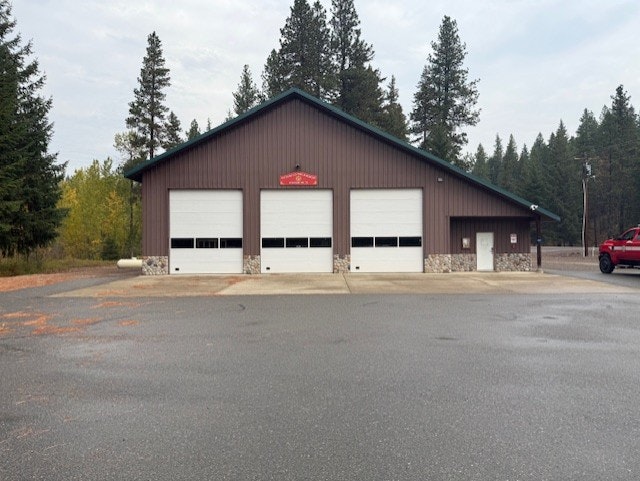 A large brown building with three garage doors and a small sign above. It’s surrounded by trees and a red vehicle is to the right.