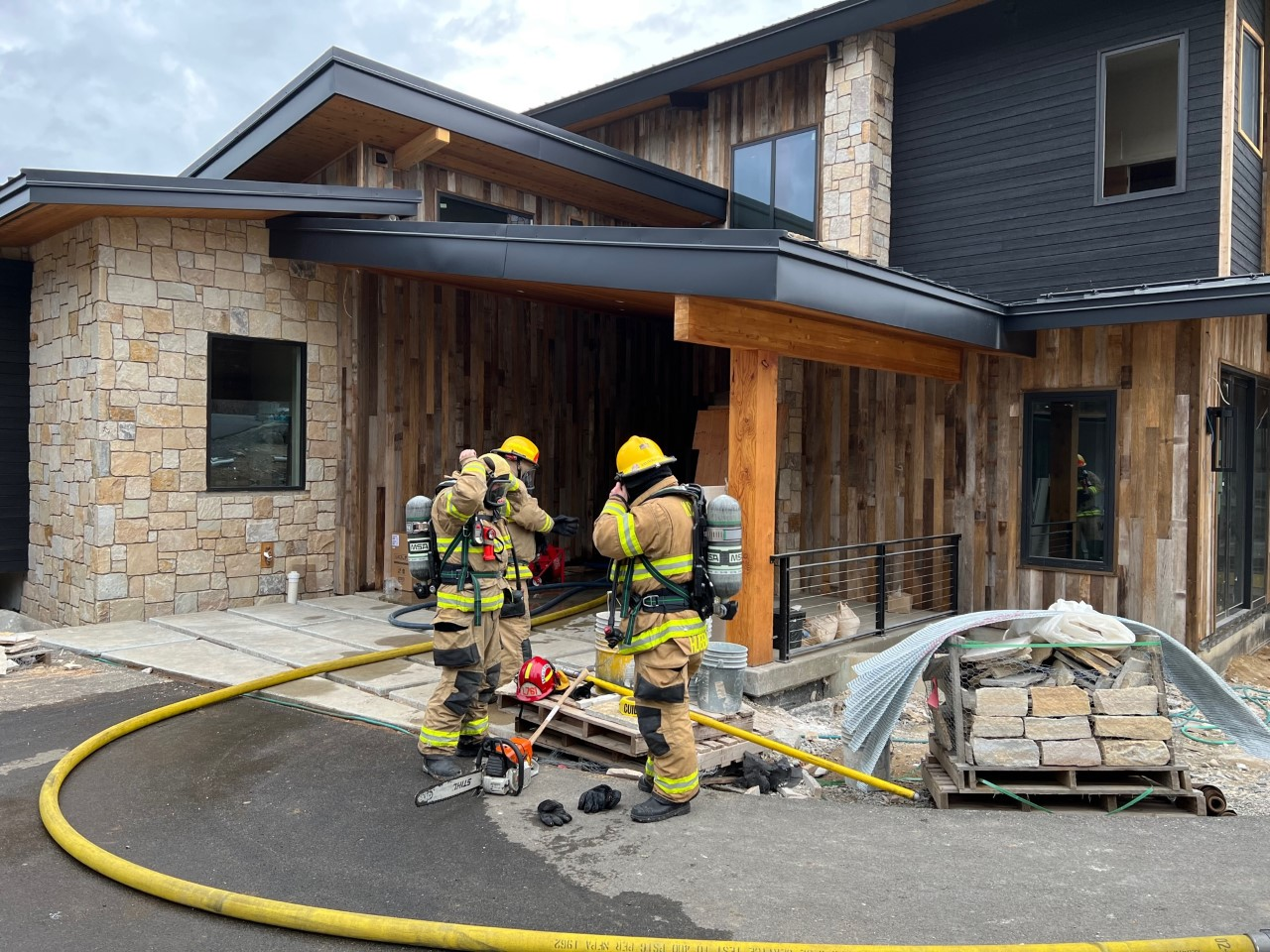Two firefighters in gear, with hoses and equipment, outside a stone and wood building.