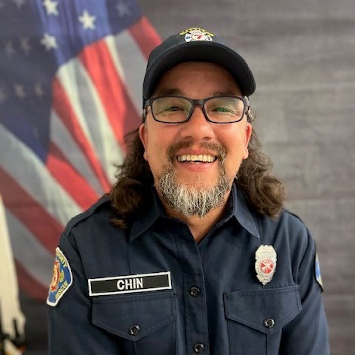 A smiling man in a uniform with badges, standing in front of an American flag backdrop.
