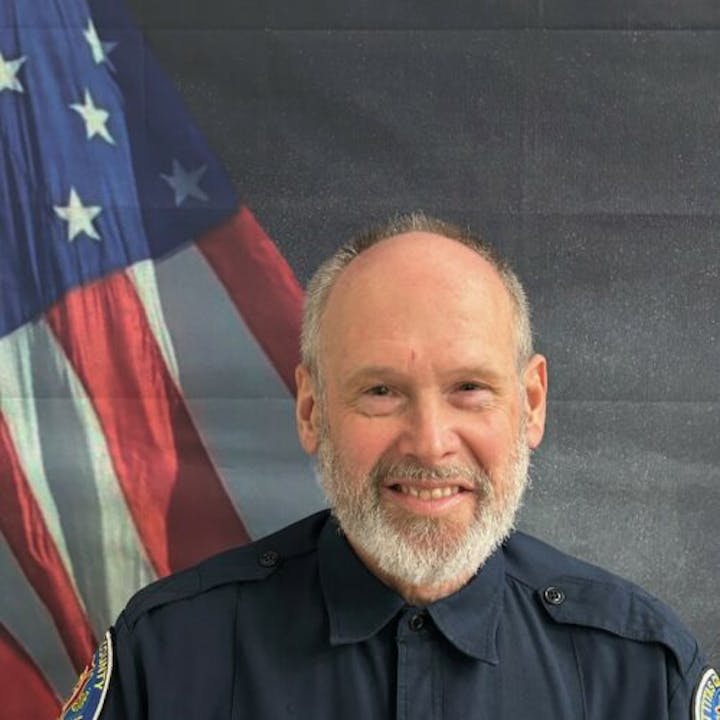 A man in a uniform stands in front of a U.S. flag backdrop, smiling.