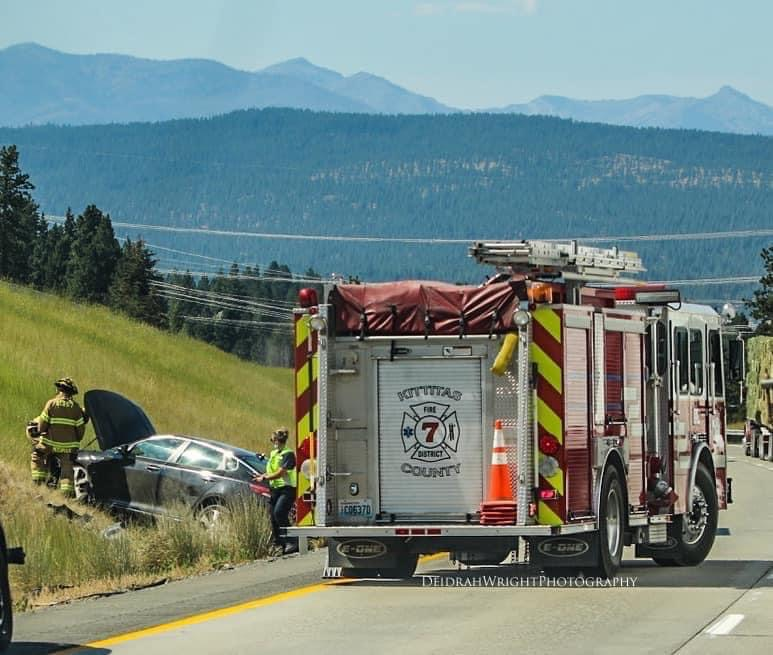 Firetruck and firefighters responding to a car off the road in a grassy area with mountains in the background.