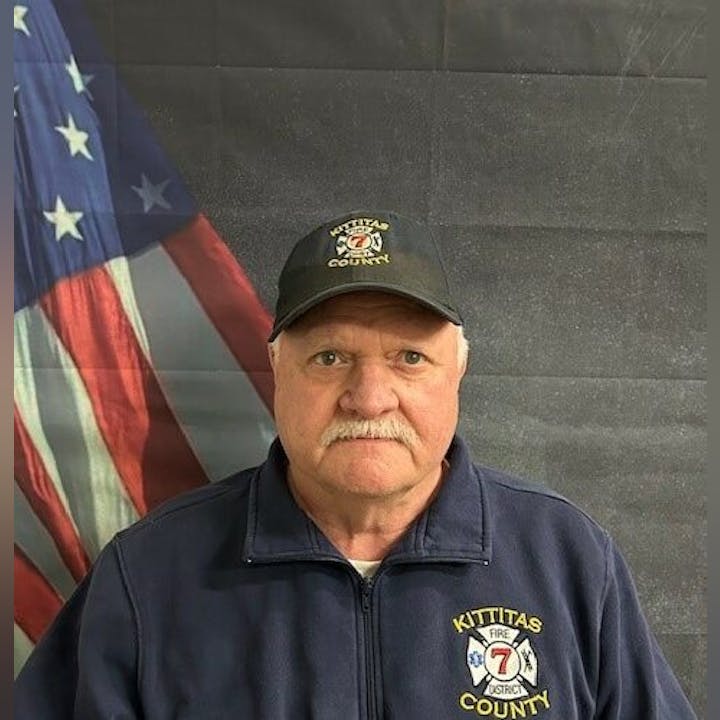 A man wearing a "Kittitas County Fire District 7" jacket and cap stands in front of an American flag backdrop.