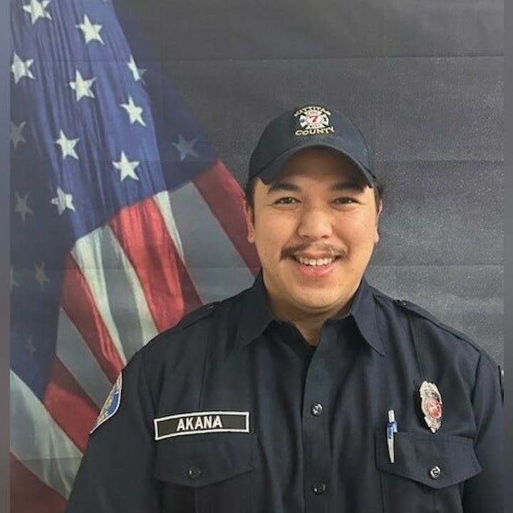 A person in a uniform stands in front of an American flag, smiling.
