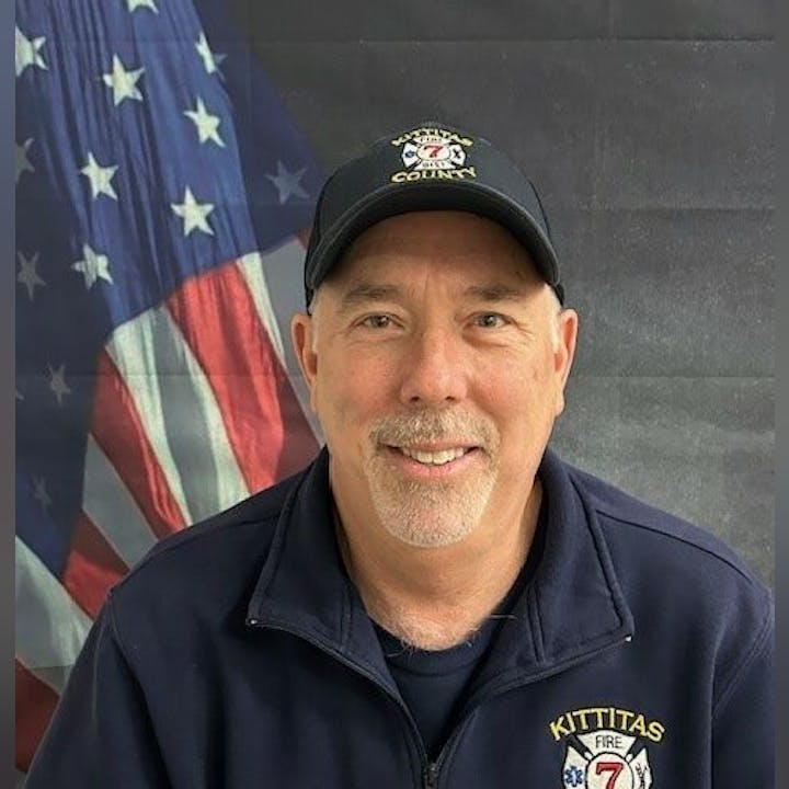 A man in a Kittitas Fire 7 jacket and cap, smiling in front of a US flag backdrop.