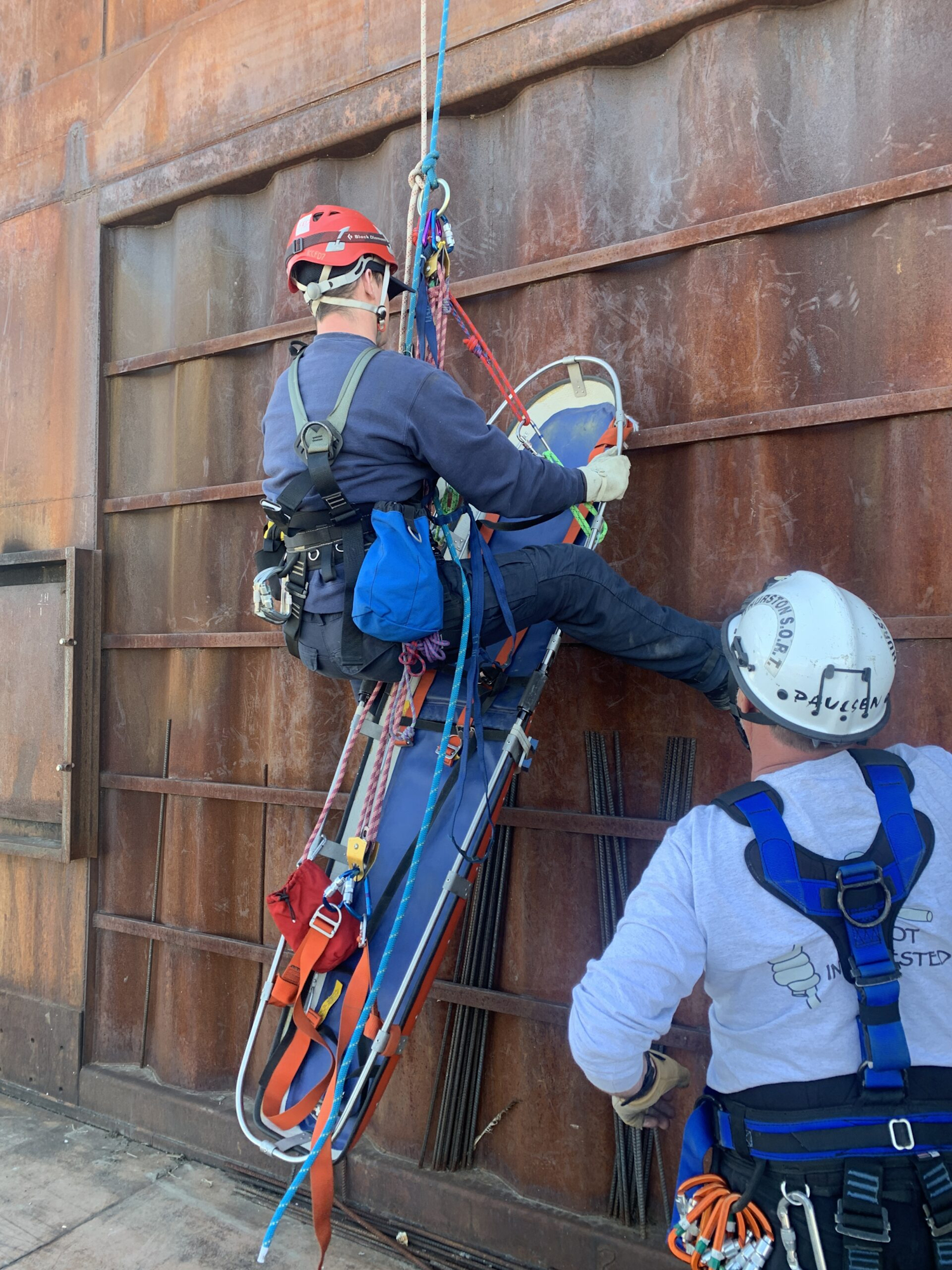 Two rescue workers in harnesses practice lowering a stretcher down a rusty industrial structure.