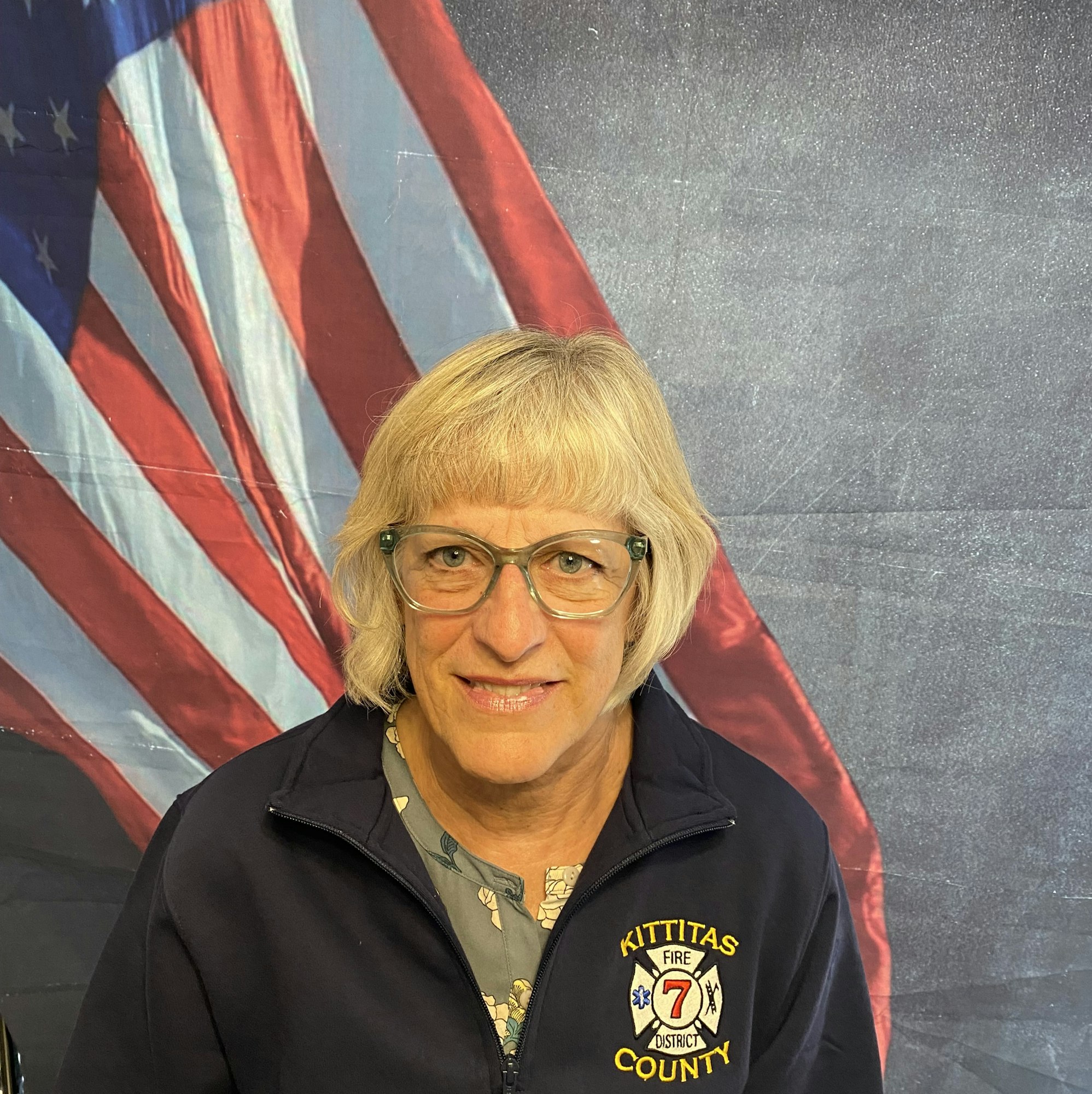 A person in a Kittitas County Fire District 7 jacket, standing in front of an American flag backdrop, smiling at the camera.
