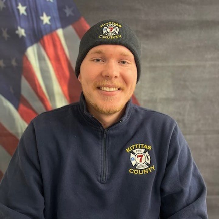 A person smiling, wearing a "Kittitas County Fire" uniform with a hat, in front of a U.S. flag backdrop.