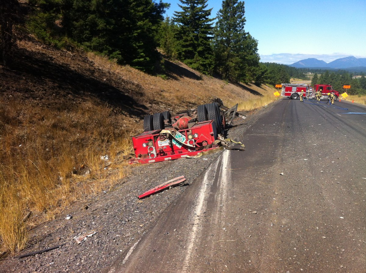 Overturned red truck on a rural road, fire trucks and firefighters in the background.