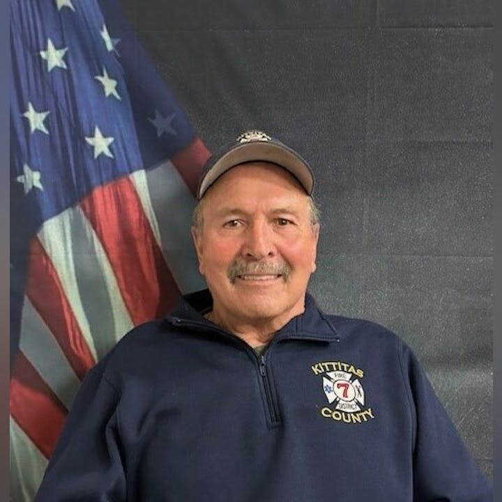 A person in a cap and "Kittitas County" jacket stands before a U.S. flag backdrop.