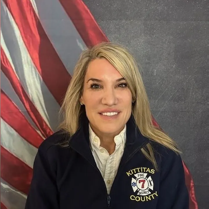 A person with long hair wearing a "Kittitas County Fire District 7" jacket, in front of an American flag backdrop.