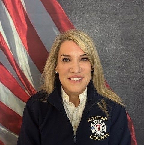A person with long hair wearing a "Kittitas County Fire District 7" jacket, in front of an American flag backdrop.