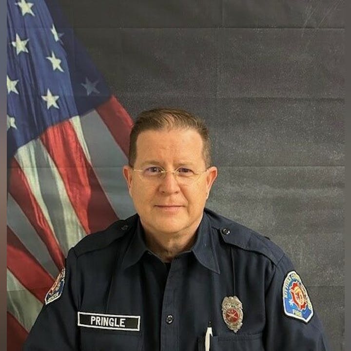 A person in a dark uniform with badges stands in front of an American flag backdrop.