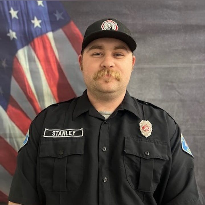 A person in a black uniform with a name badge, standing in front of a flag.