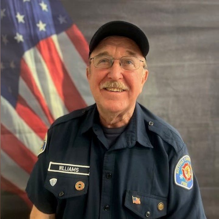A man in a uniform with "Williams" on the badge stands in front of an American flag, smiling.
