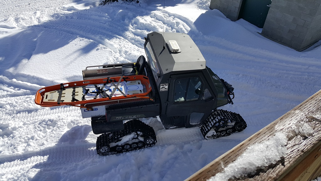 A tracked vehicle on snow carrying an orange rescue sled.