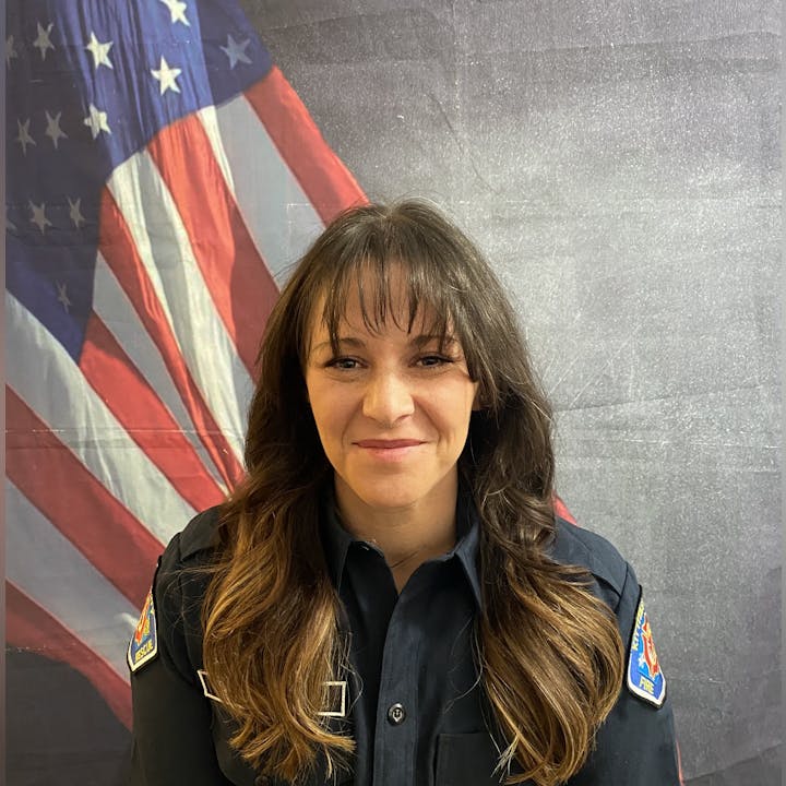 A person in a dark uniform stands in front of an American flag backdrop.