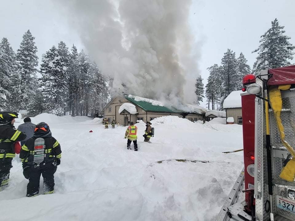 Firefighters battle a snowy house fire, smoke rises from the roof, surrounded by snow-covered trees.