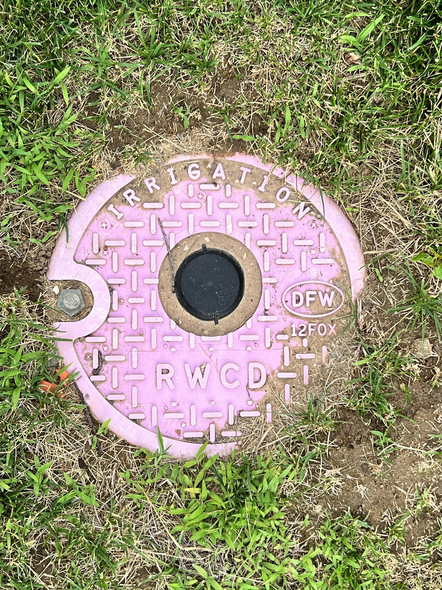 A purple irrigation cover surrounded by grass with text: "IRRIGATION RWCD DFW 12FOX."