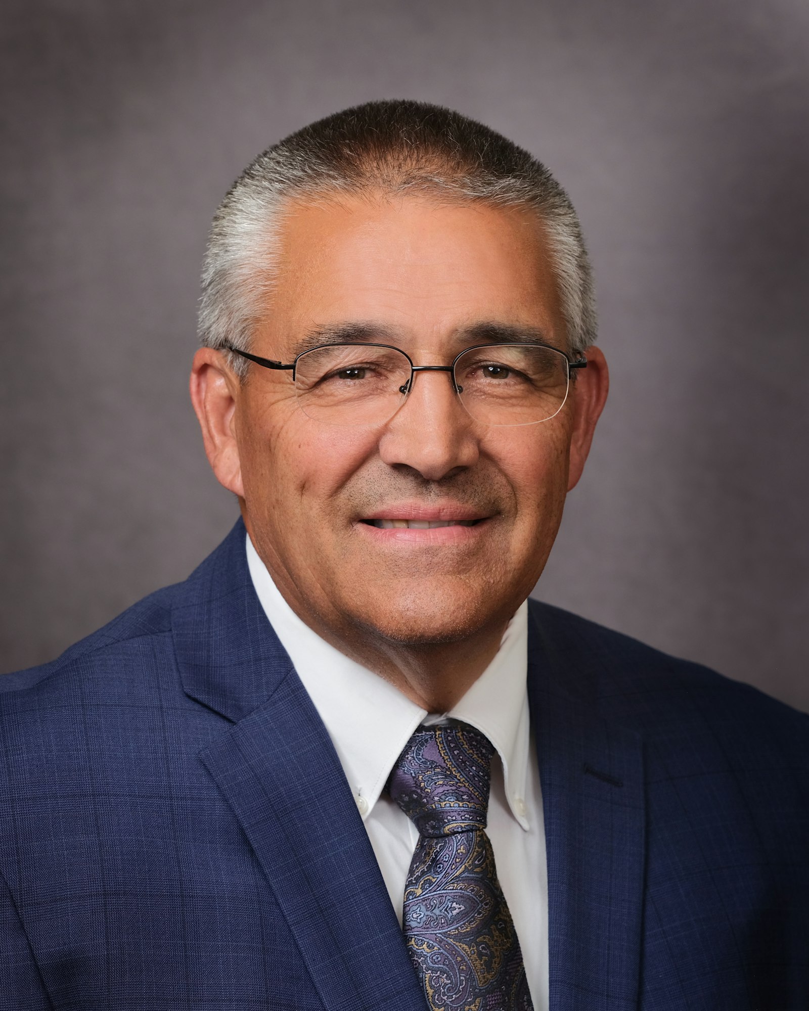 A man wearing glasses, a blue suit, and a paisley tie, smiling against a gray background.