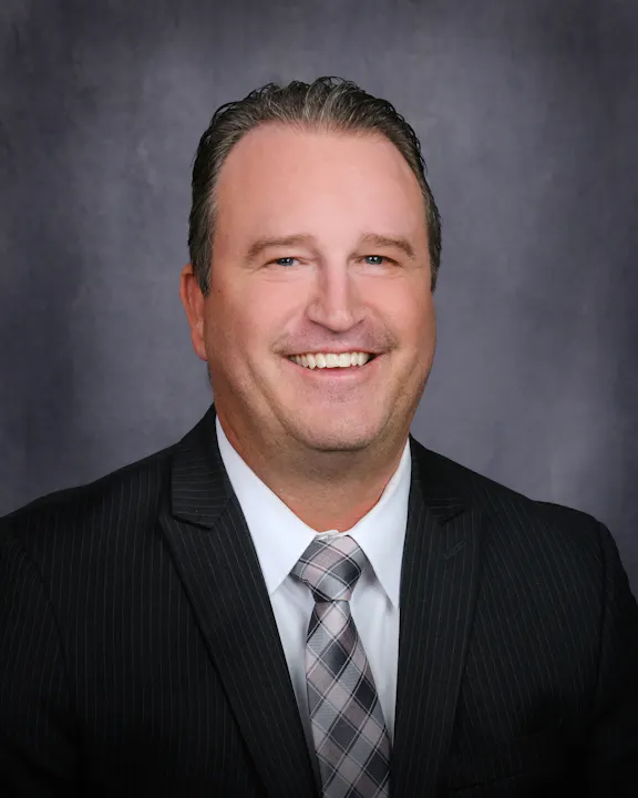 The image features a smiling man in business attire, with a gray background. He's wearing a suit with a tie and has short hair.