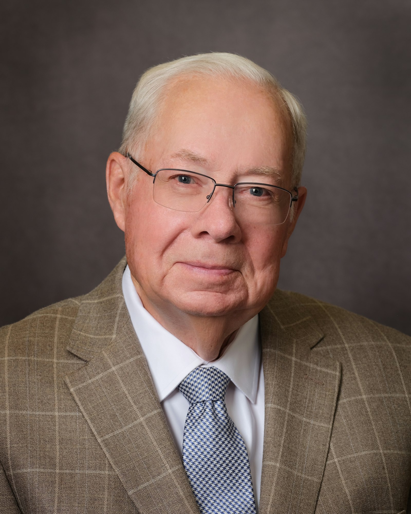 A man with glasses in a checkered suit, white shirt, and patterned tie, against a plain background.