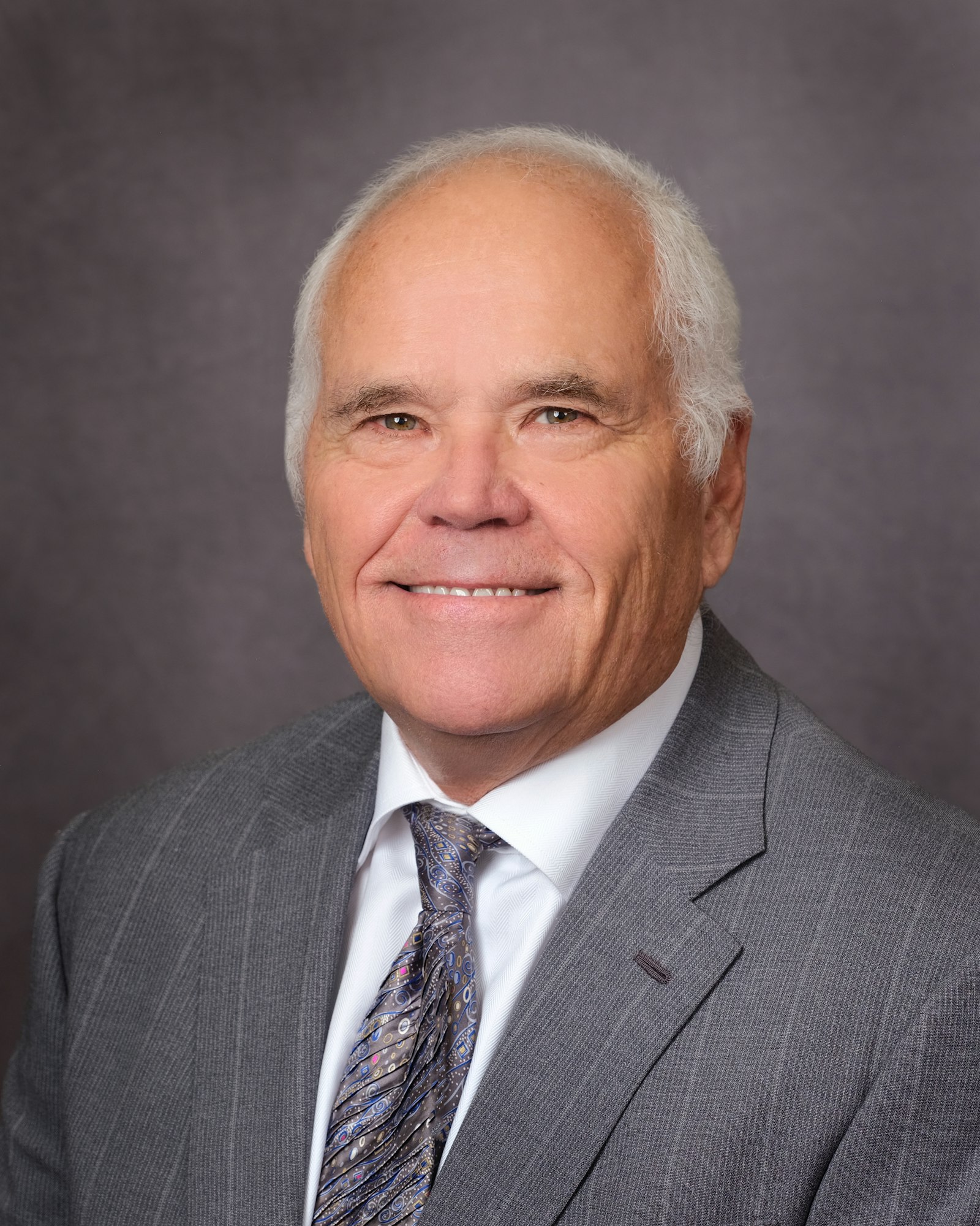 A person in a gray suit and tie smiling in front of a plain background.