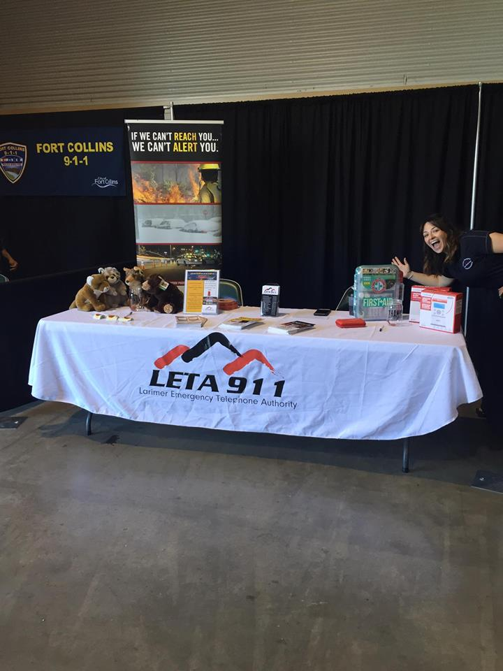 Exhibition booth with information banners, a smiling person, promotional items, and stuffed toys on the table.