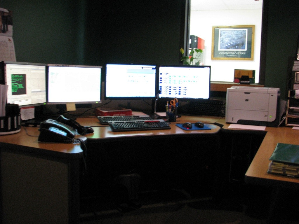 Office desk with multiple monitors, phone, keyboard, mouse, printer, and office supplies.