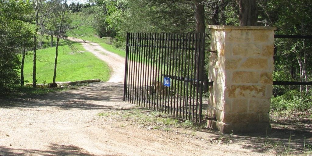 A metal gate with a stone pillar on a rural dirt road, trees, grass, and a blue address sign.