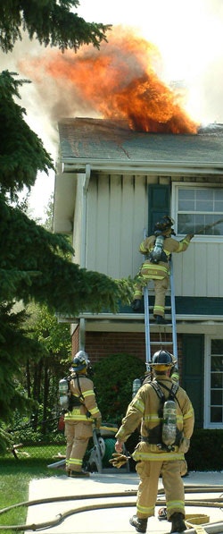 Firefighters at a house fire, with smoke and flames on the roof. One climbs a ladder, others stand on the ground.