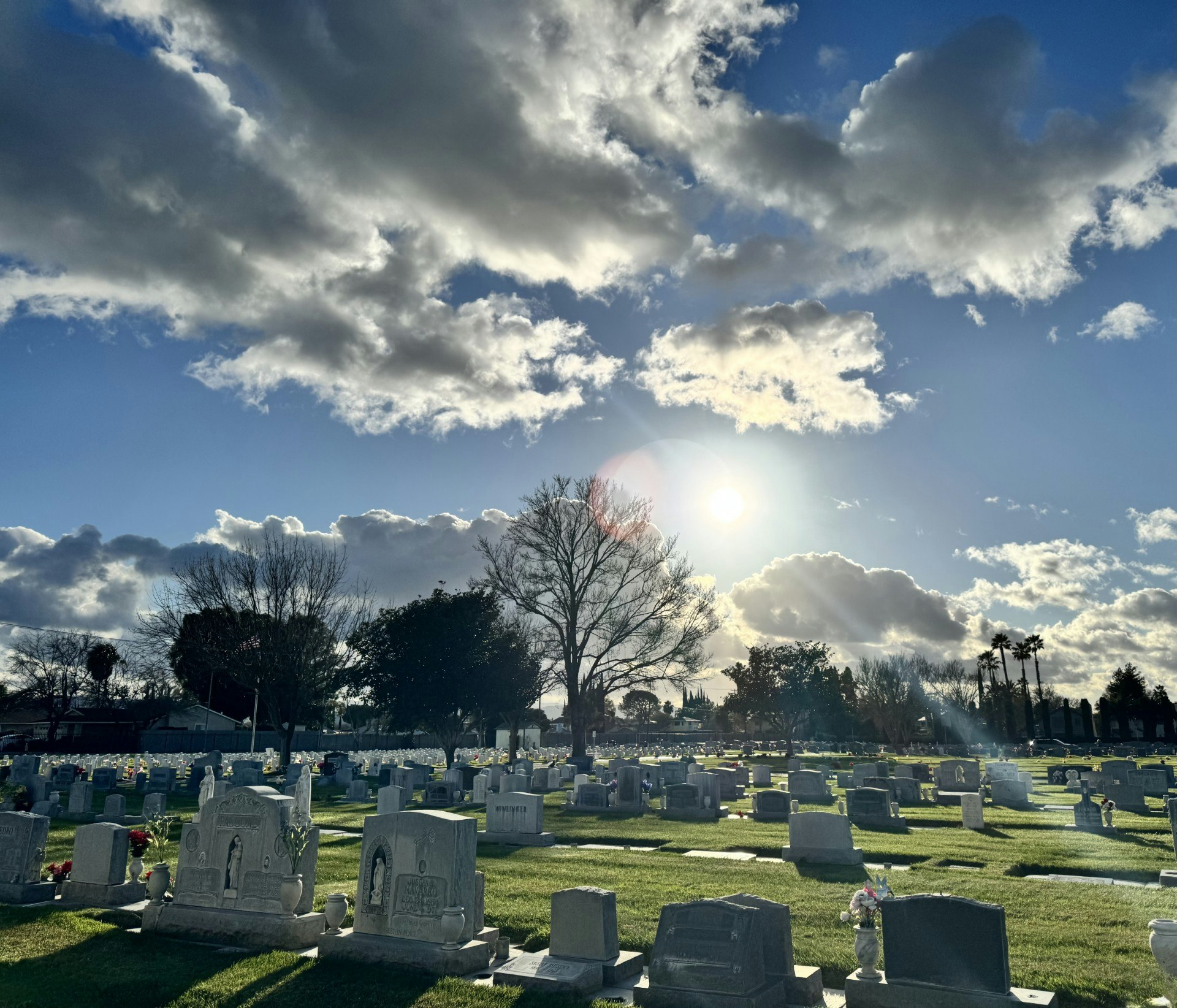 A serene cemetery scene with gravestones, green grass, and a bright sun shining amidst a cloudy sky.