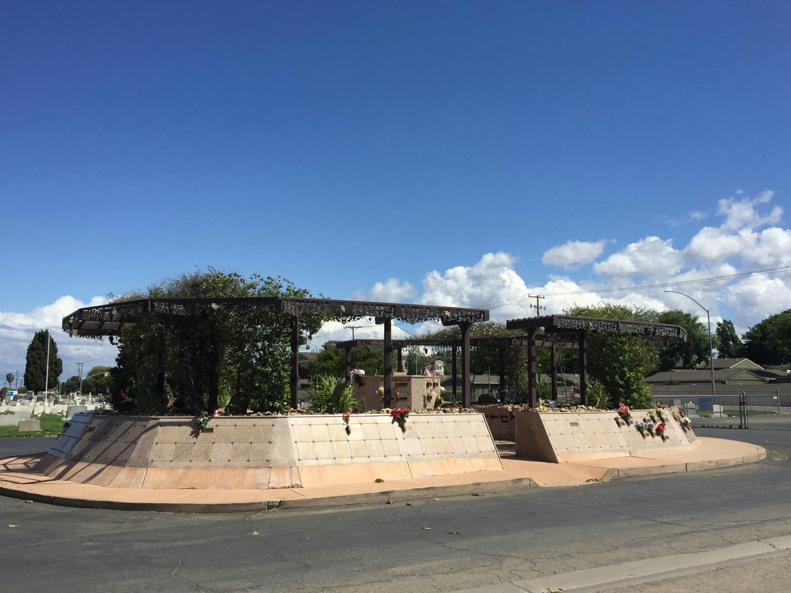 A landscaped structure with a metal canopy and plants, surrounded by a road, under a blue sky with clouds.