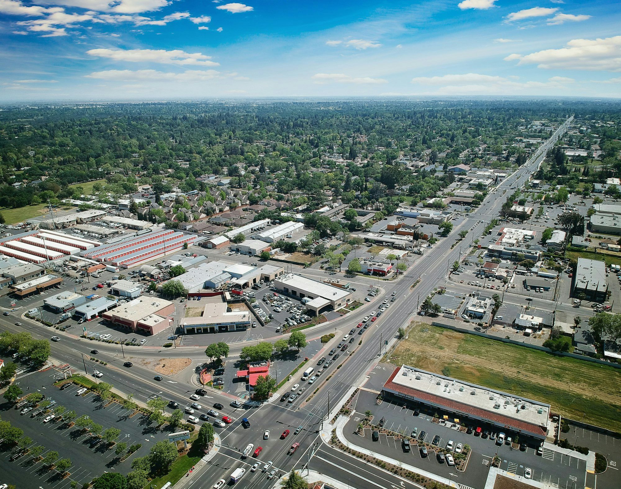 Aerial view of a cityscape with roads, buildings, parking lots, and greenery under a blue sky.