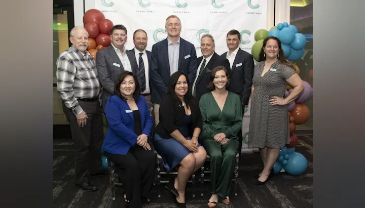 A group photo of ten individuals posing together at an event, with a backdrop of balloons and a branding banner.