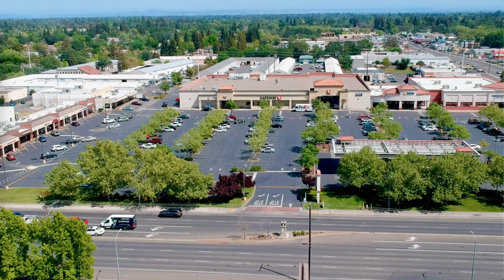 Aerial view of a shopping center with a supermarket, parking lot, and surrounding trees. Busy road in the foreground.